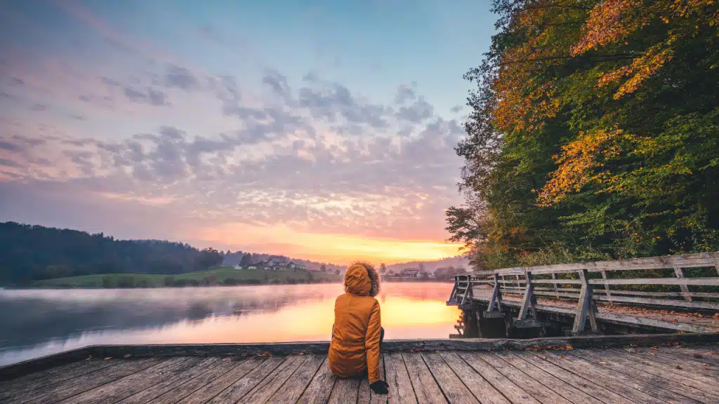 Une personne de dos avec un manteau est assise au bord d'un ponton de bois avec vue sur un lac et des habitations au coucher du soleil